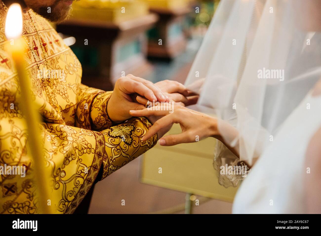 Vista del sacerdote messo su anello sulla mano di spose, posa in chiesa cristiana durante la cerimonia di nozze, candela bruciante in primo piano Foto Stock