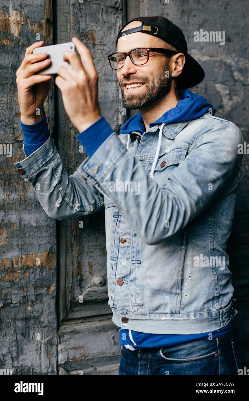 Maschio eccitato con barba e baffi che indossano una camicia trendy in denim con gancio e jeans che guarda felicemente nella fotocamera del suo smartphone facendo selfie. Secondo Foto Stock