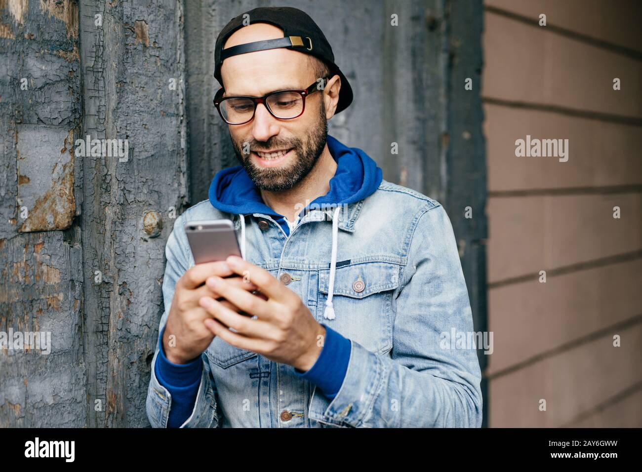 Ritratto orizzontale di bell'uomo elegante bearded in denim anorak indossando grande occhiali da vista che tiene smartphone utilizzando Internet gratuito, controllare e-mail, wa Foto Stock