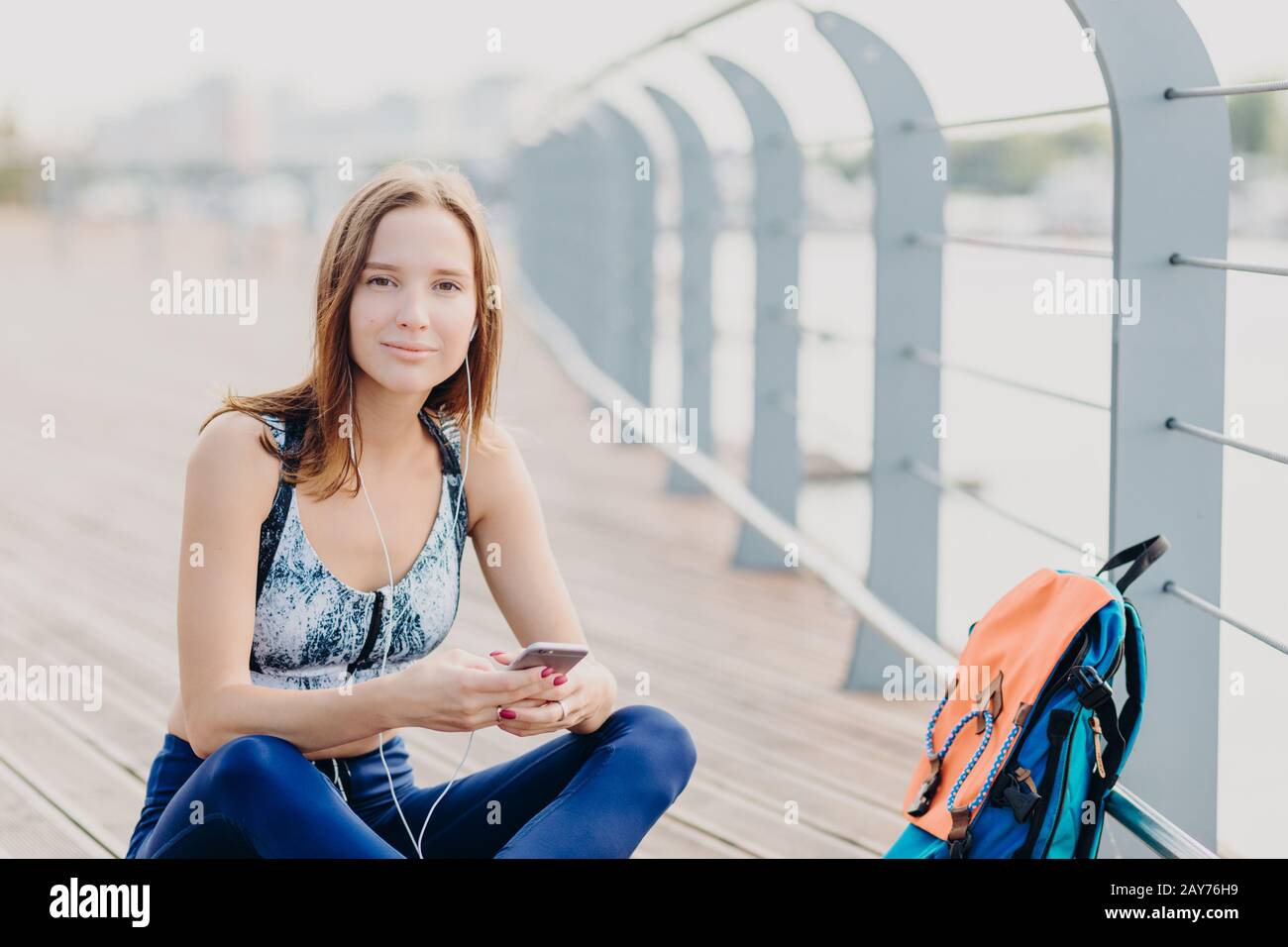 Foto di attraente giovane sportswoman felice con pelle sana, va dentro per lo sport regolarmente, siede le gambe incrociate con il telefono mobile, gode favorito Foto Stock