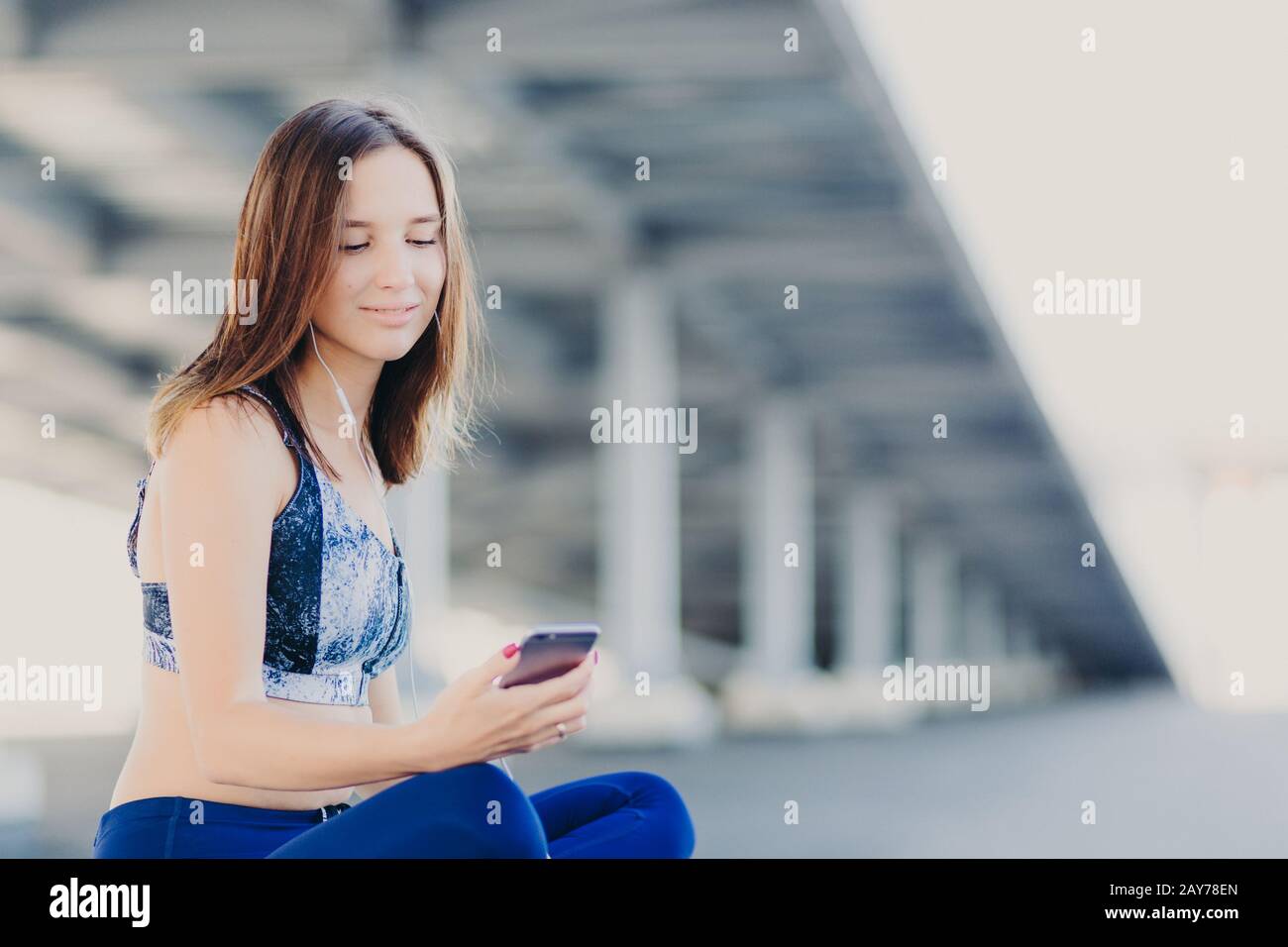 Scatto orizzontale della donna graziosa graziosa con l'apparenza piacevole, ascolta la radio, tiene il telefono delle cellule moderno, usa gli auricolari e la connessione wifi libera, dres Foto Stock