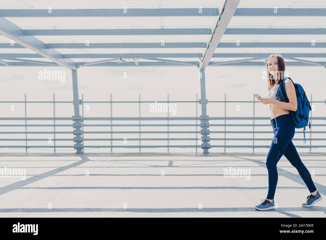 Positivo sottile ragazza fitness in corso di formazione, essere fotografato in movimenti, porta borsa, tiene cellulare moderno, utilizza auricolari aggiornati, stand o Foto Stock