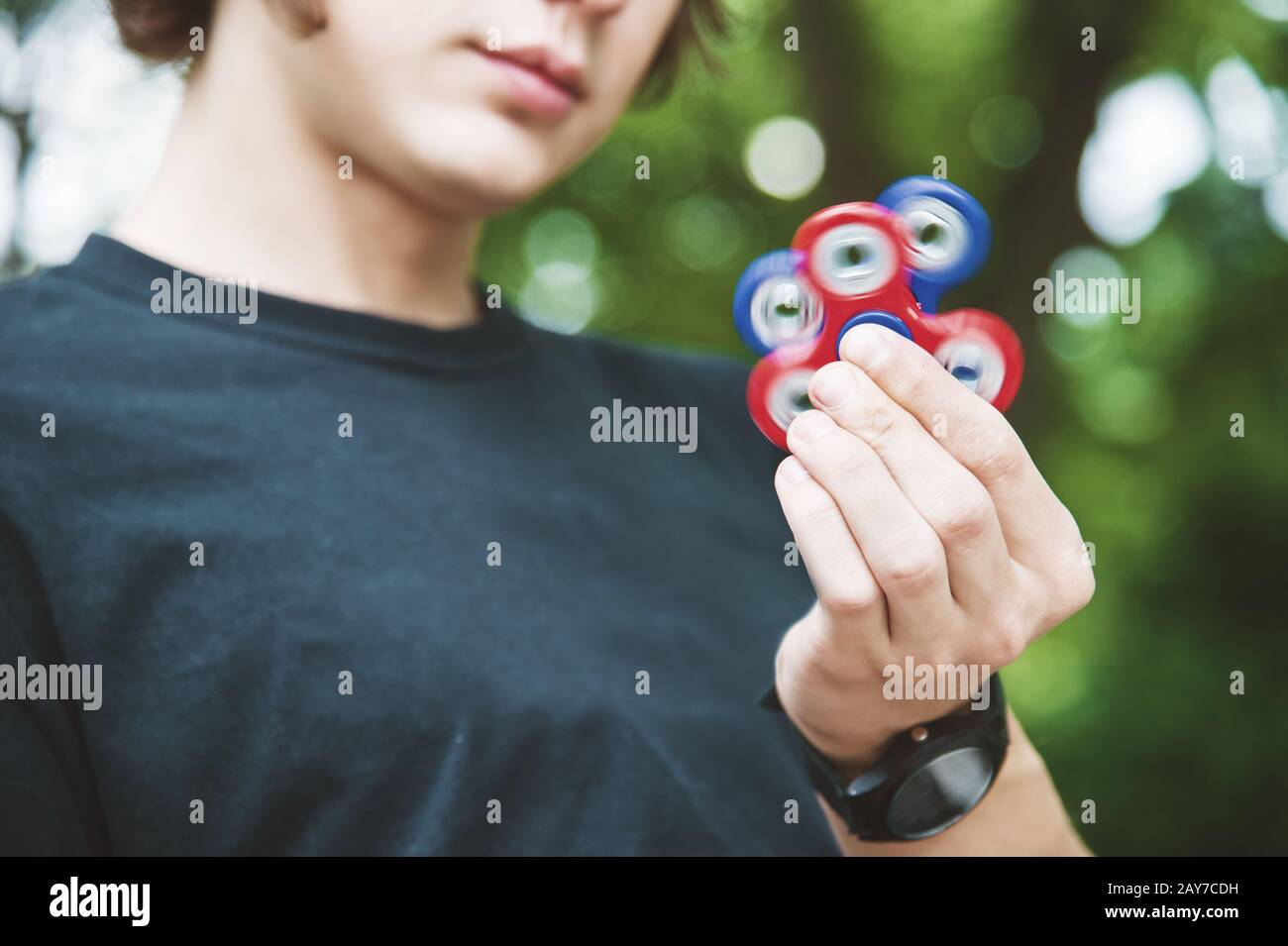 Un hipster dai capelli lunghi in un cappuccio si siede su una panchina e fa girare un fidget-spinner Foto Stock