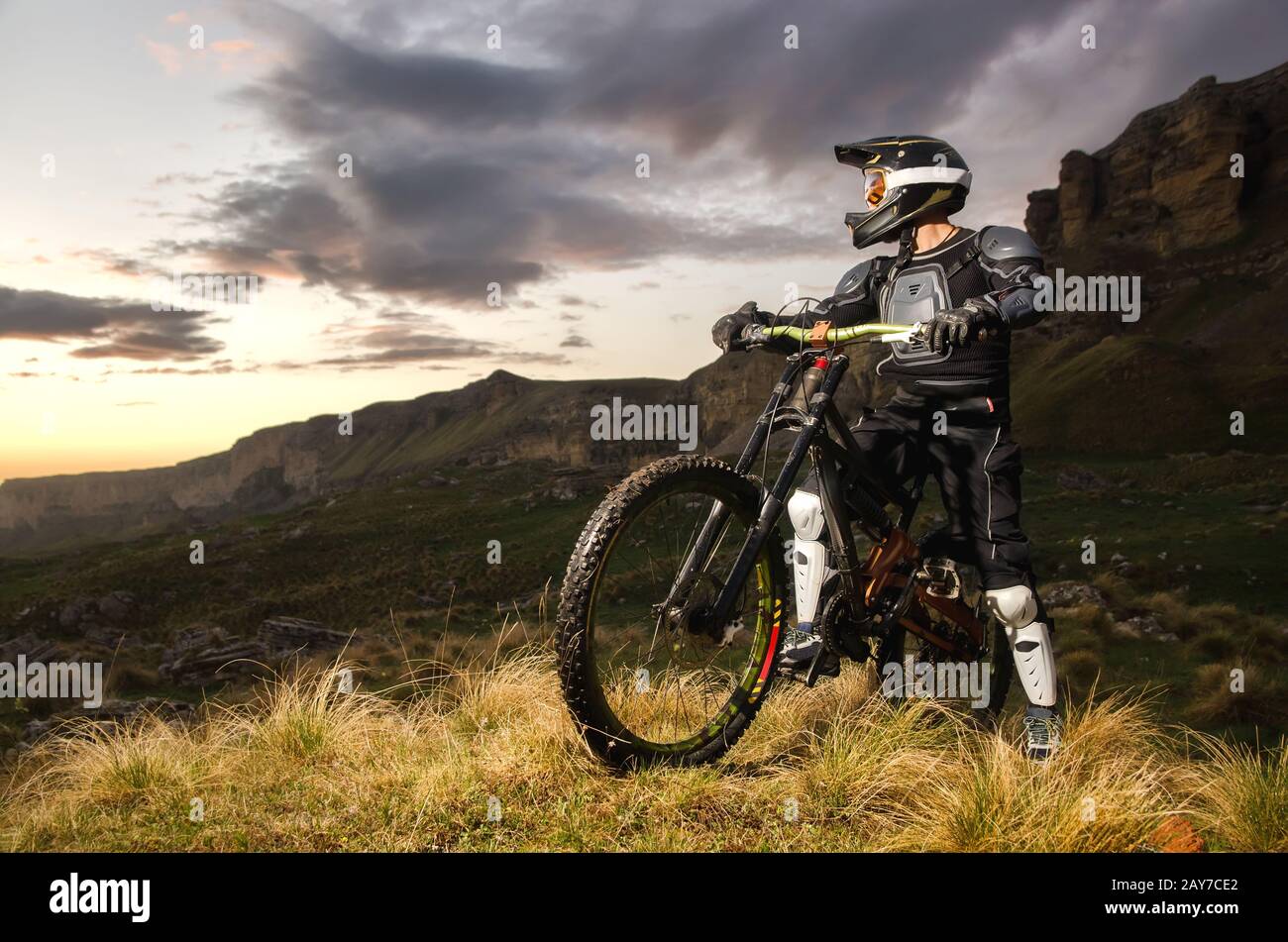 Il pilota in piena protezione su una mountain bike si alza e guarda il tramonto sullo sfondo delle rocce Foto Stock
