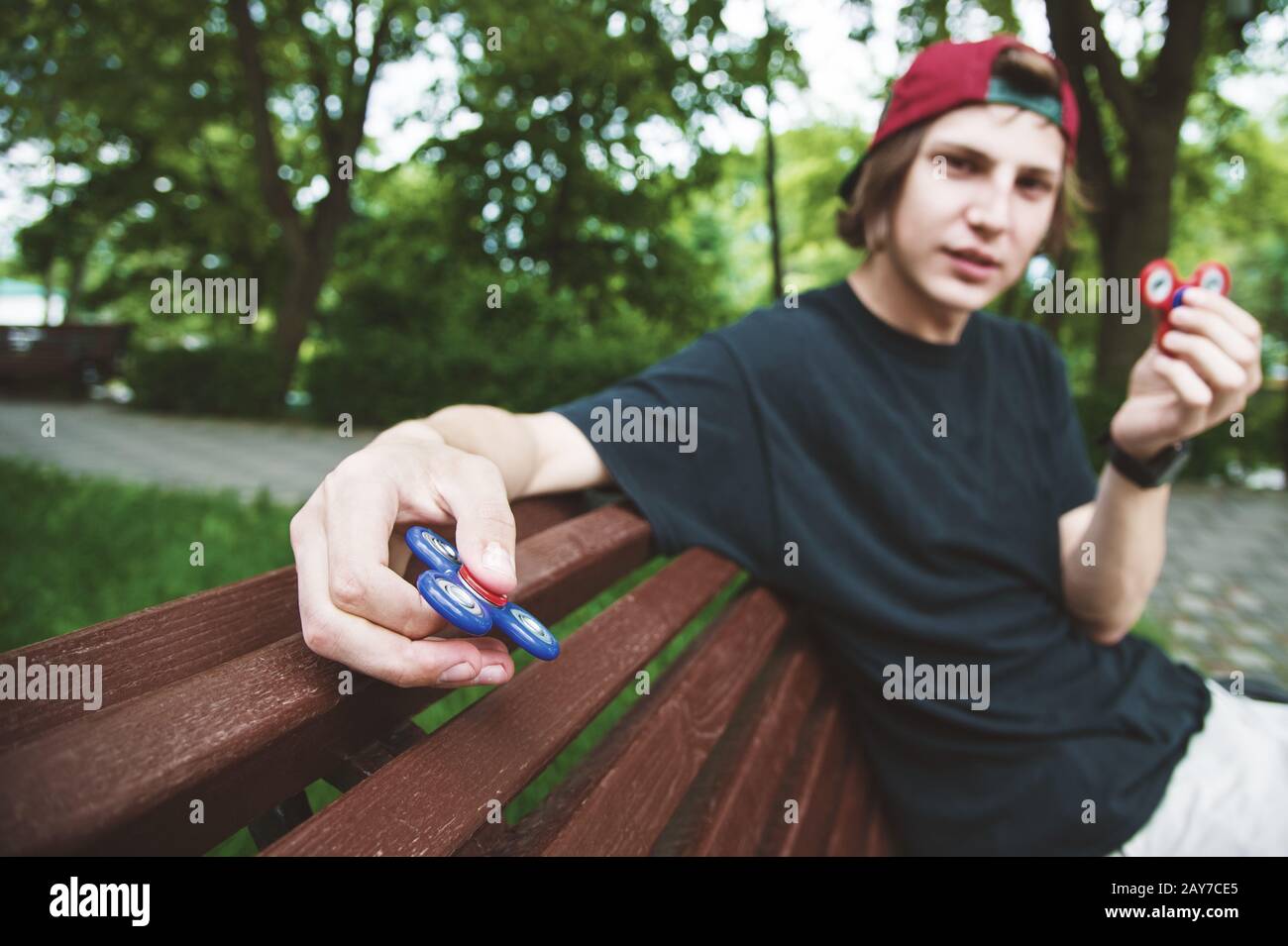 Un hipster dai capelli lunghi in un cappuccio si siede su una panchina e fa girare un fidget-spinner Foto Stock