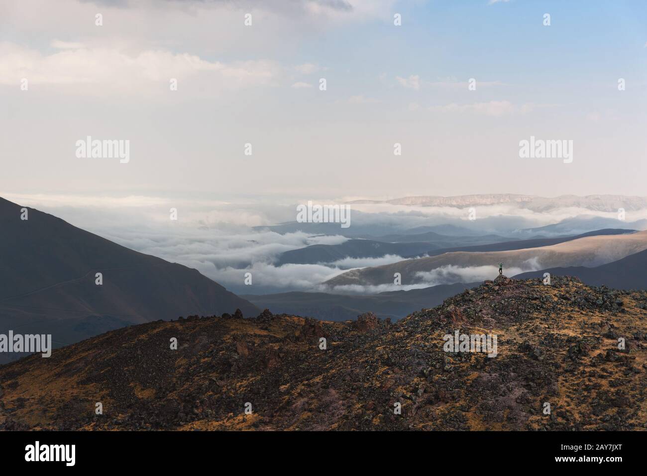 Escursionista il vincitore si trova sul bordo di una roccia con bastoni sollevati di fronte a una valle coperta da nuvole nel Caucaso Foto Stock