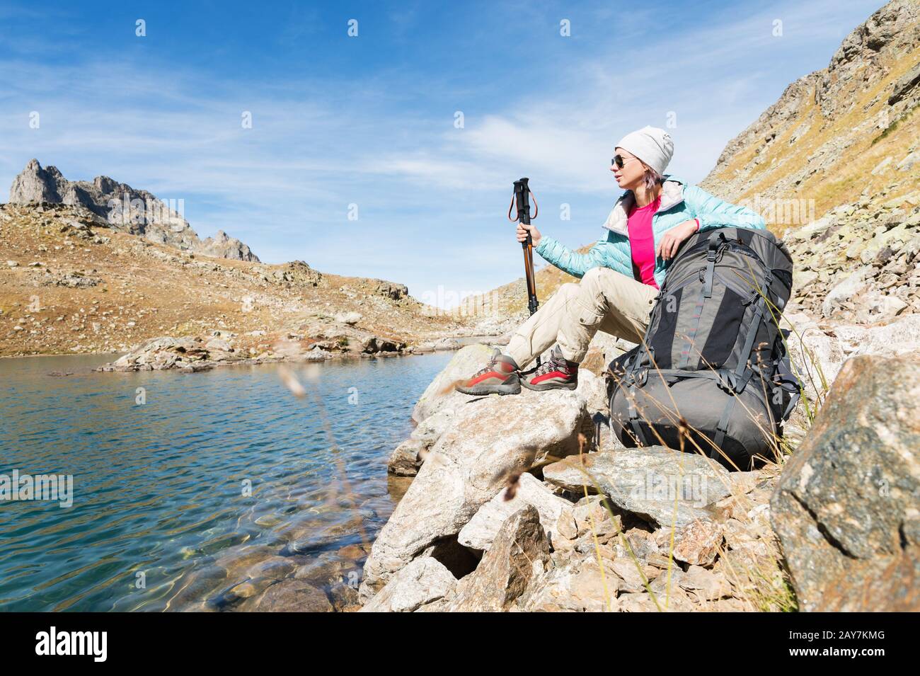 Una ragazza turistica che indossa occhiali da sole giacca e cappello con uno zaino e attrezzatura di montagna con manici per il tracking in ha Foto Stock