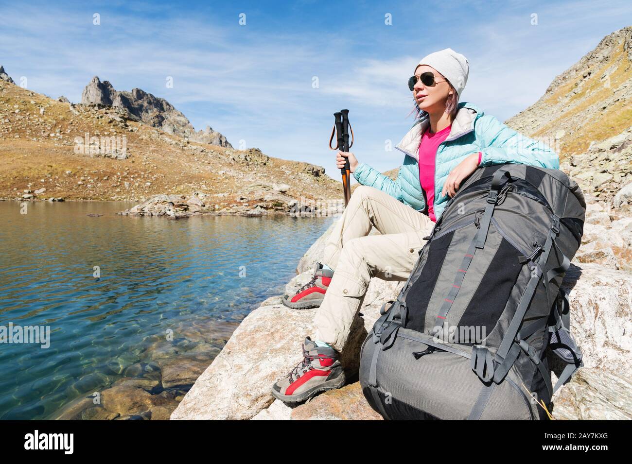 Una ragazza turistica che indossa occhiali da sole giacca e cappello con uno zaino e attrezzatura di montagna con manici per il tracking in ha Foto Stock