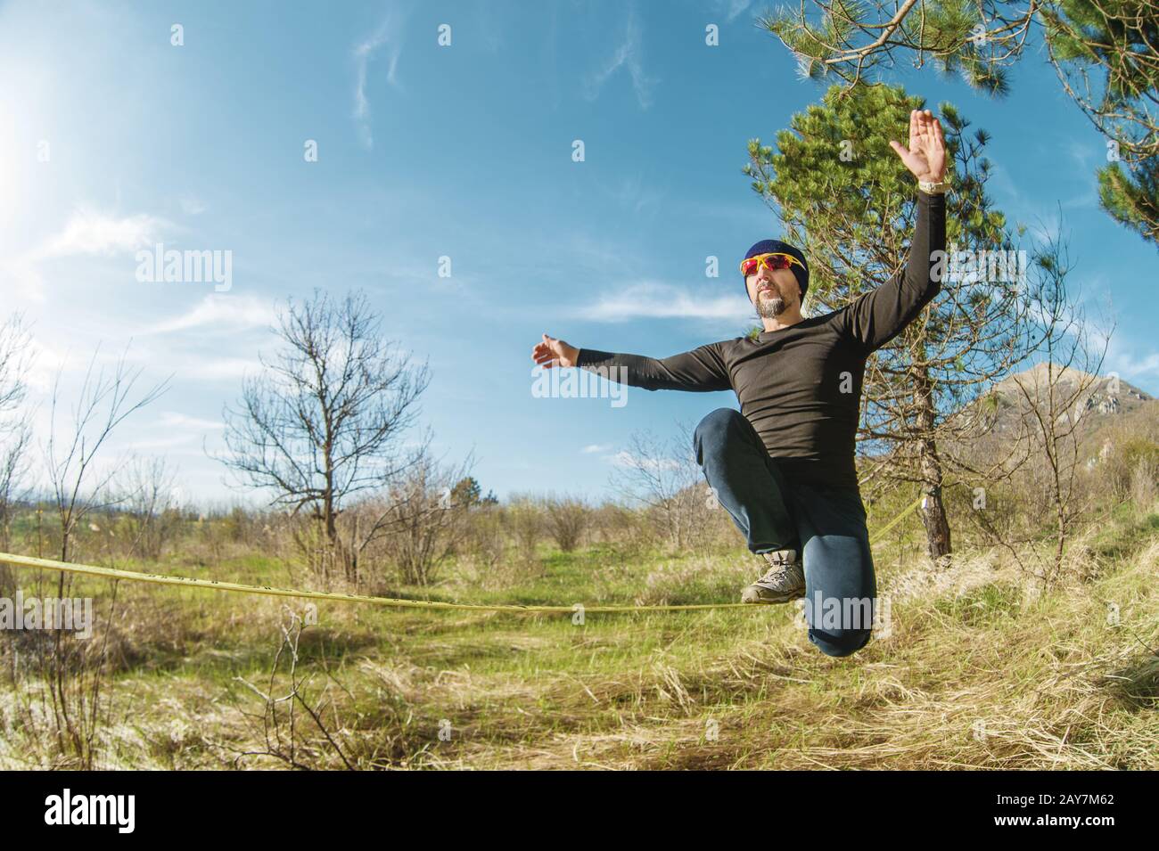 Un uomo all'età di sedersi in occhiali con un cappello e scarpe da ginnastica sul slackline, cattura l'equilibrio e gode la vita sulla natura i. Foto Stock