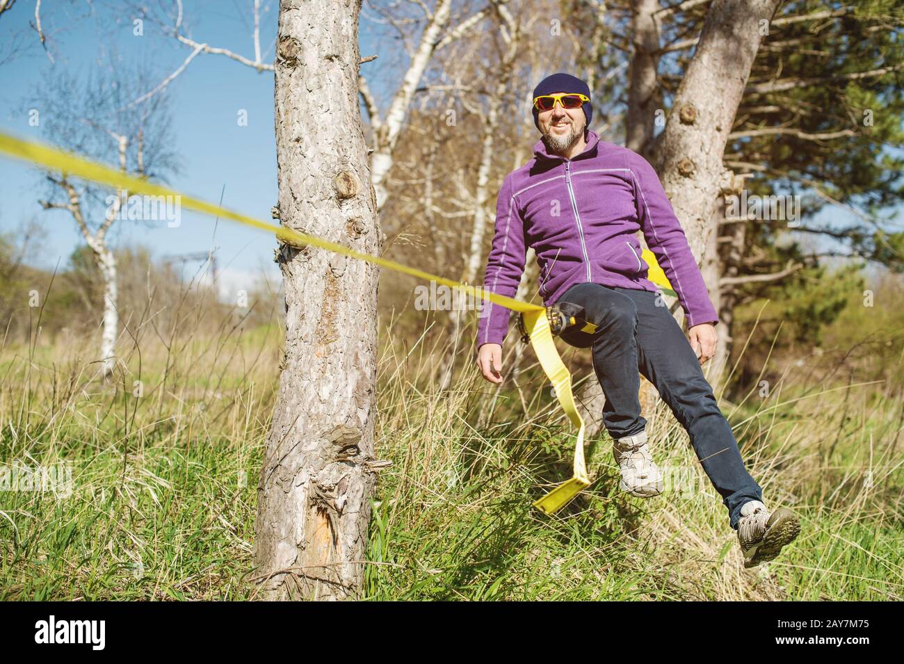 Un uomo all'età di sedersi in occhiali con un cappello e scarpe da ginnastica sul slackline, cattura l'equilibrio e gode la vita sulla natura i. Foto Stock