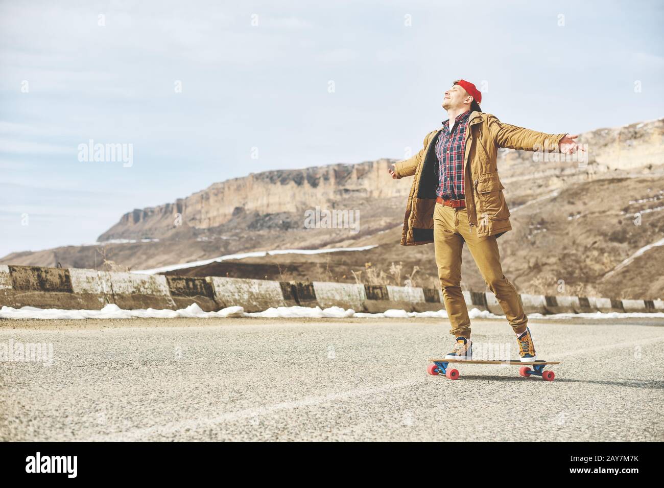 Giovane ragazzo felice elegante in un cappello e pantaloni joggers rotolare lungo una strada di montagna su un longboard, godendo la vita Foto Stock