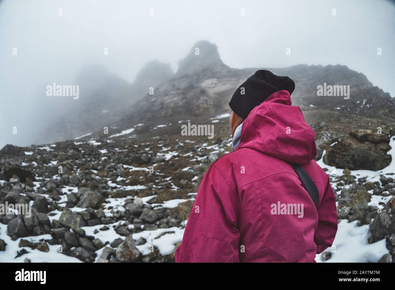 Una giovane ragazza da sola sulle montagne caucasiche guarda le rocce ai piedi delle quali si trova la neve. Le rocce avvolgono nubi nubi nebbiose Foto Stock