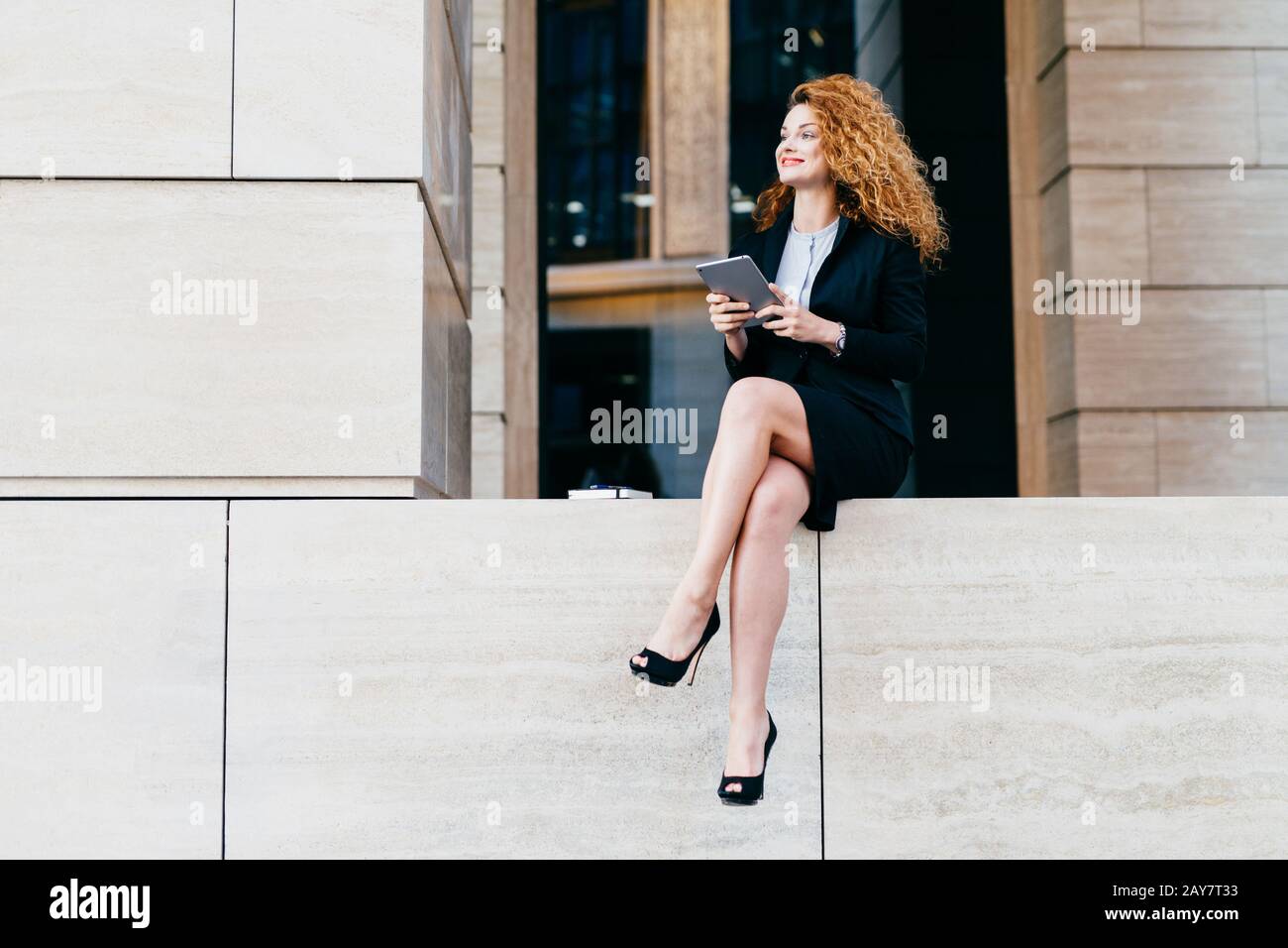 Ritratto di elegante donna splendida in abito nero e scarpe alte-heeled con gambe sottili lunghe, seduta con tablet moderno, guardando da parte con felice Foto Stock