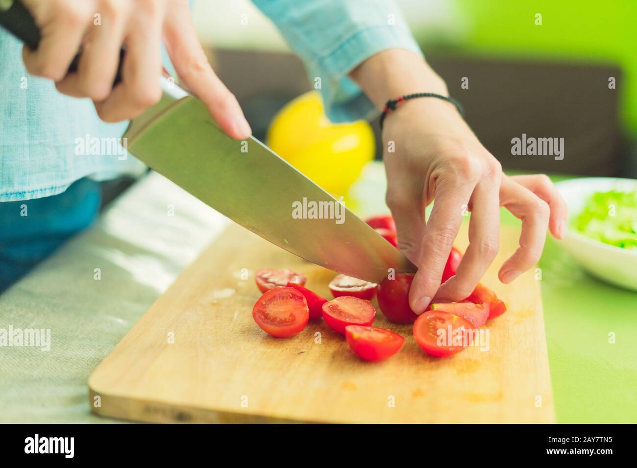 Le mani di una giovane ragazza tritate i pomodori ciliegini su un tagliere di legno su un tavolo verde in un ambiente domestico Foto Stock