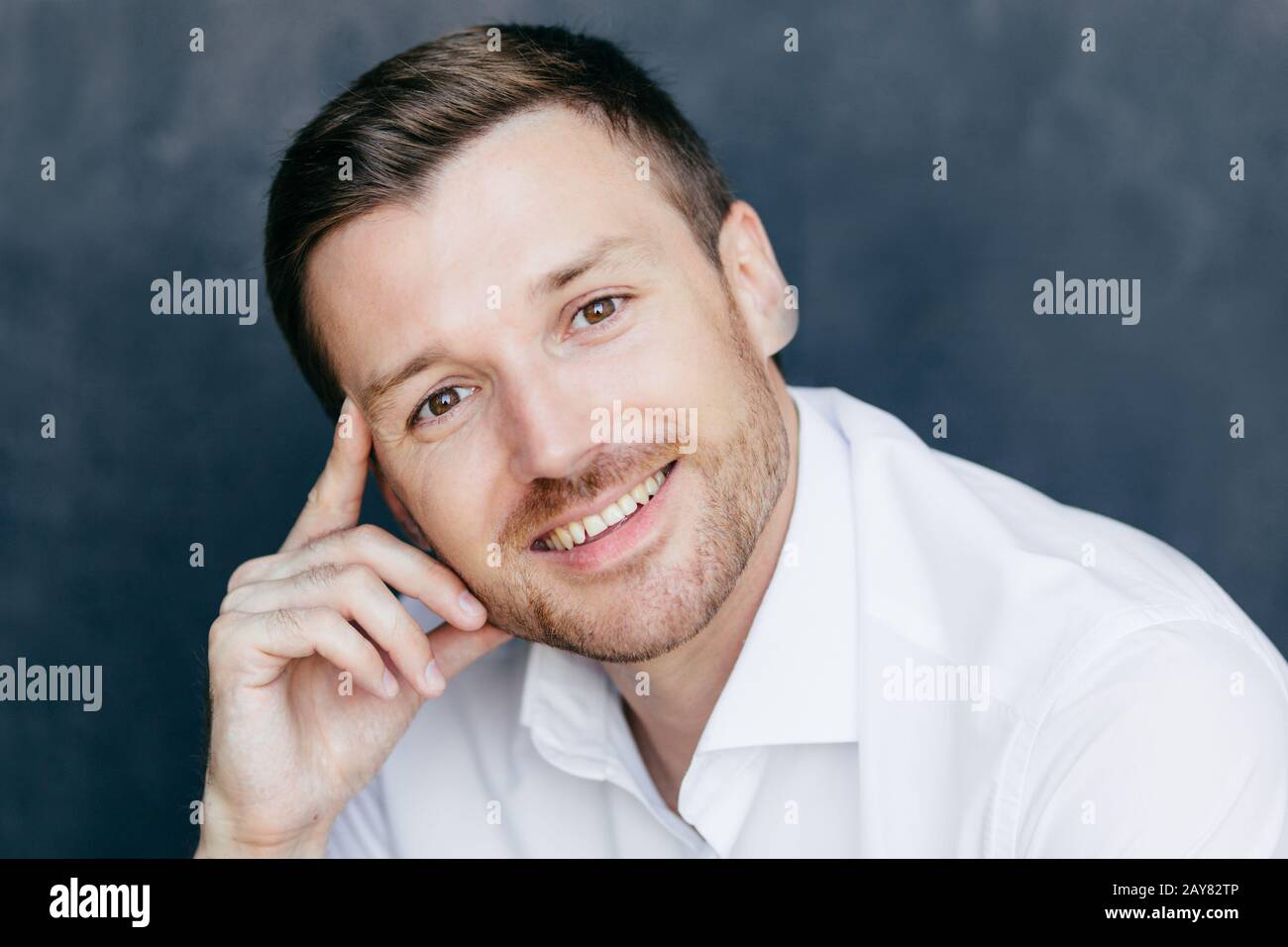 Allegro uomo dipendente ha espressione gioiosa, tiene dito sul tempio, gioisce iniziare la vacanza, vestito in formale camicia bianca, pone contro la ba scuro Foto Stock