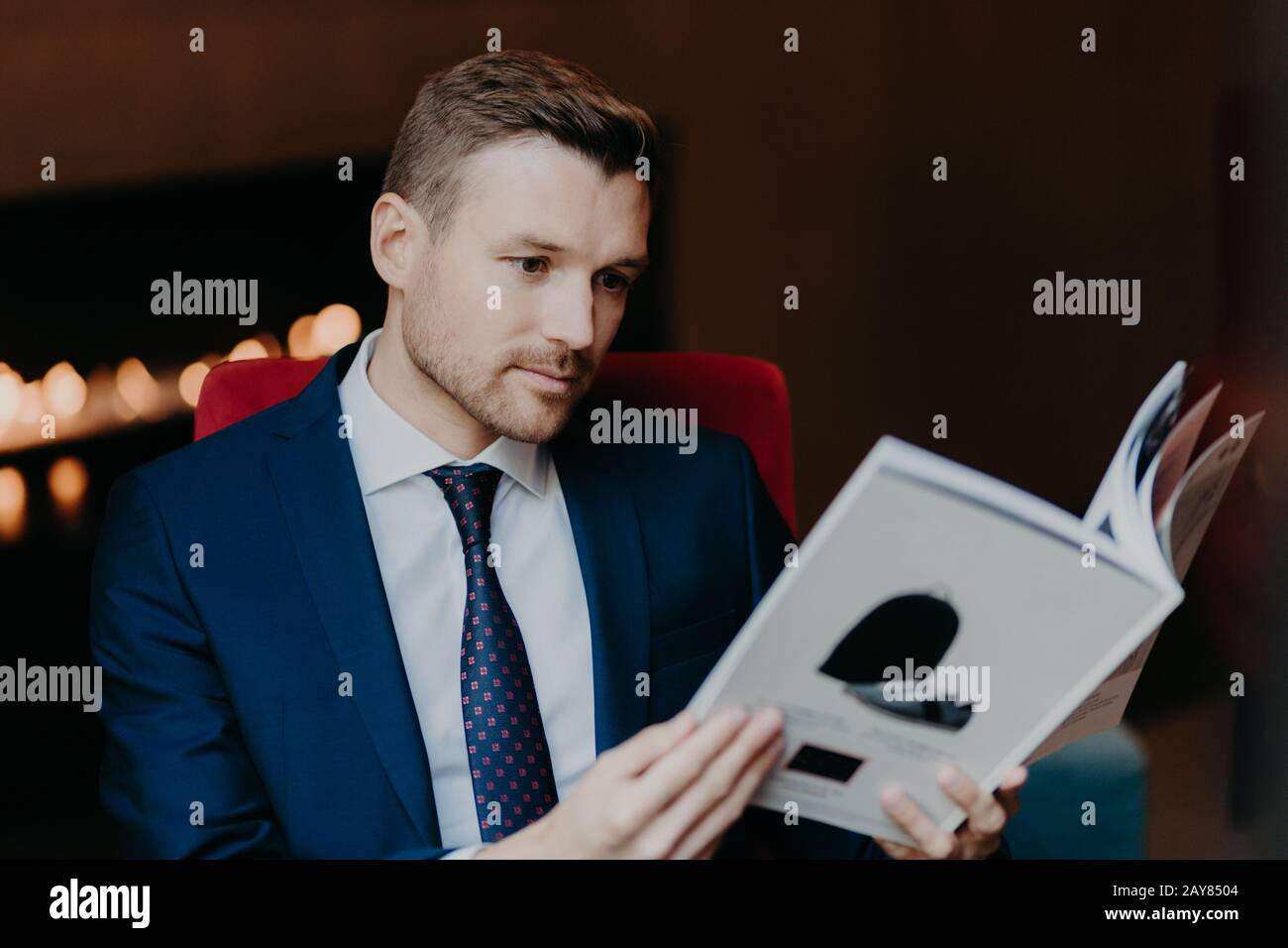 Concentrato giovane boss ha stoppia, essendo ben vestito, legge interessante rivista mentre si siede in caffetteria e attende per ordine, legge interessanti informazioni Foto Stock