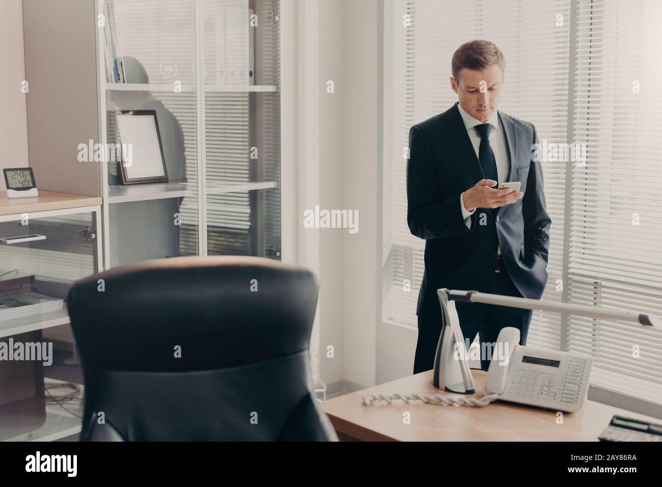 Bel giovane manager maschile in abbigliamento aziendale, pagine web browses e chat online con smartphone, ha concentrato il look, pone in ufficio moderno Foto Stock