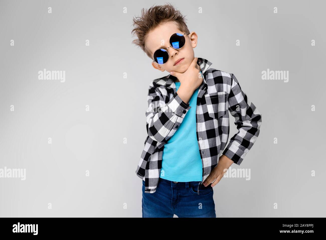 Un bel ragazzo in una camicia in plaid, camicia blu e jeans si erge su uno sfondo grigio. Il ragazzo indossa occhiali rotondi. Capelli rossi Foto Stock