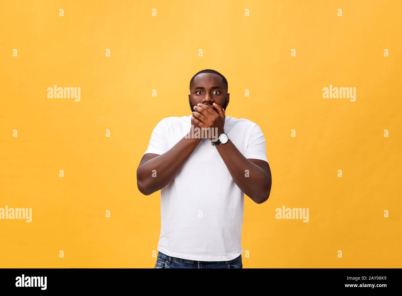 Stupito giovane afro-americano hippster che indossa la t-shirt bianca che tiene le mani nel gesto sorpreso, mantenendo la bocca aperta, guardando Foto Stock