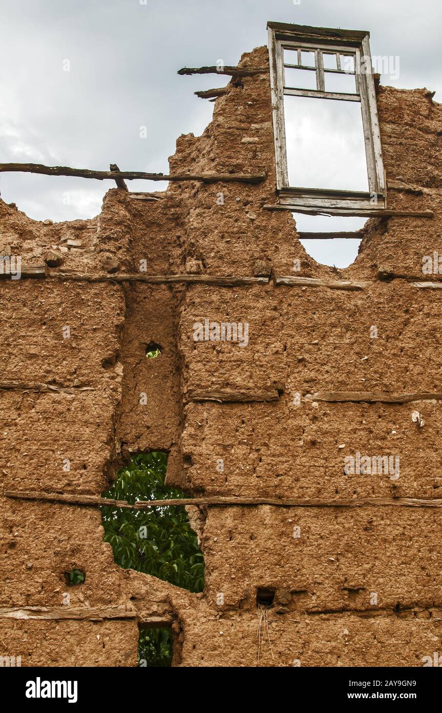 Muro di mattoni con intonaco di argilla adobe e telaio di legno rotto della finestra è rimasto di vecchia casa rurale in rovina Foto Stock