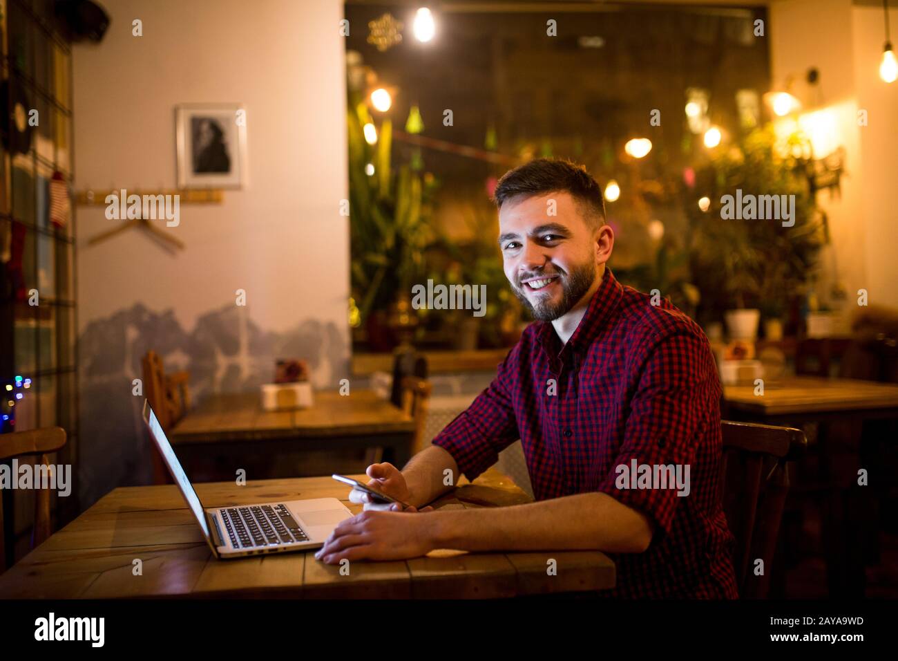 Un giovane bell'uomo caucasico con barba e un sorriso toothy in camicia rossa a scacchi sta lavorando dietro il laptop seduto a linguetta di legno Foto Stock