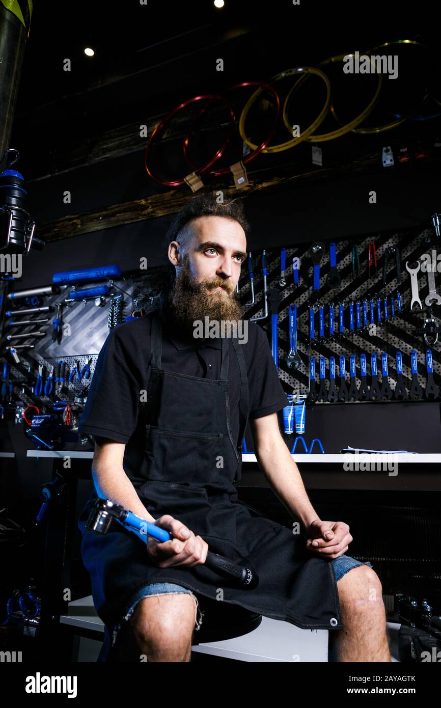 Ritratto di piccola impresa proprietario di giovane uomo con barba. Ragazzo di bicicletta meccanico officina lavoratore seduto con l'attrezzo in mano Foto Stock