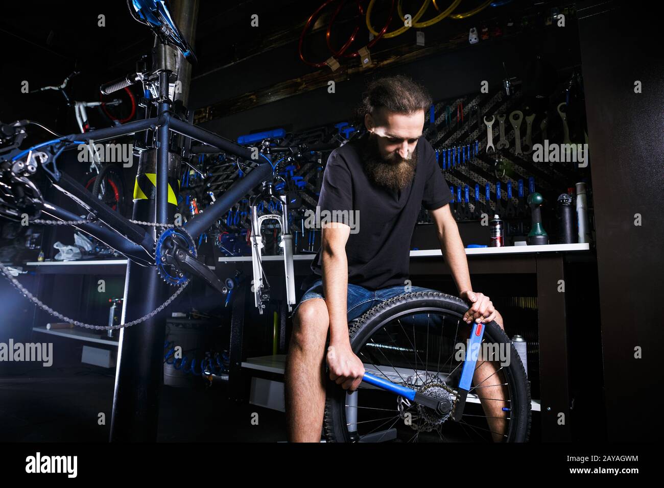 Giovane barbuto di stile, con capelli lunghi uomo meccanico caucasico in un laboratorio di biciclette utilizza uno strumento per la riparazione Delle Cassette o Foto Stock