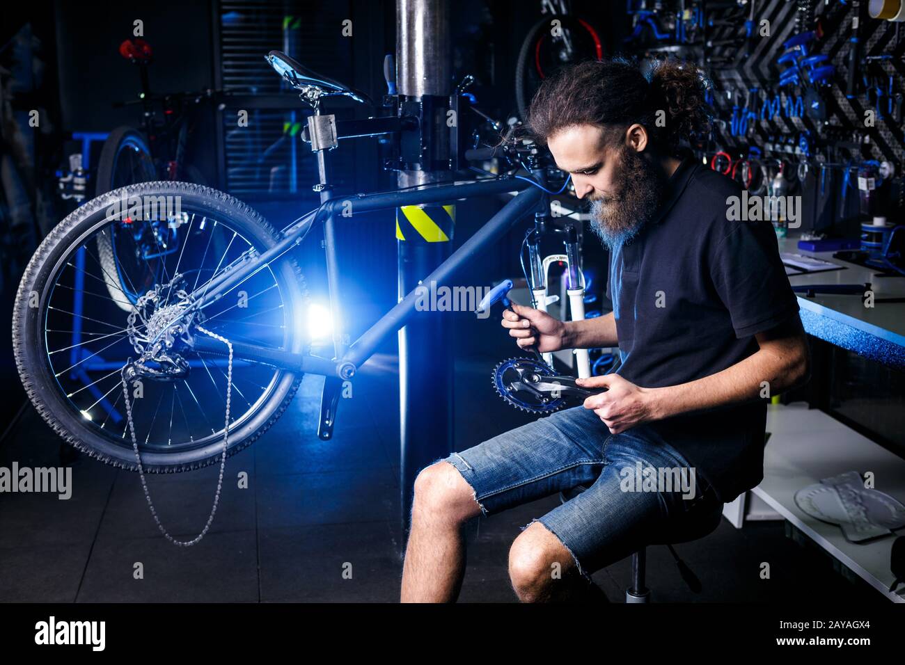 Giovane barbuto di stile, con capelli lunghi uomo meccanico caucasico in un laboratorio di biciclette utilizza un attrezzo per la riparazione Di Guarniture Foto Stock