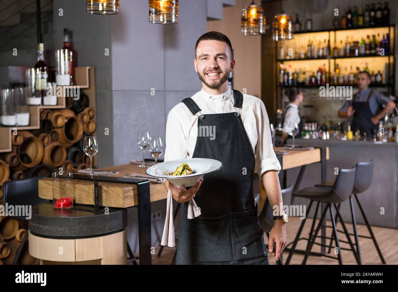 Un bel ragazzo giovane con una barba vestita in un grembiule in un ristorante e con un piatto bianco con una falda. Contro Th Foto Stock