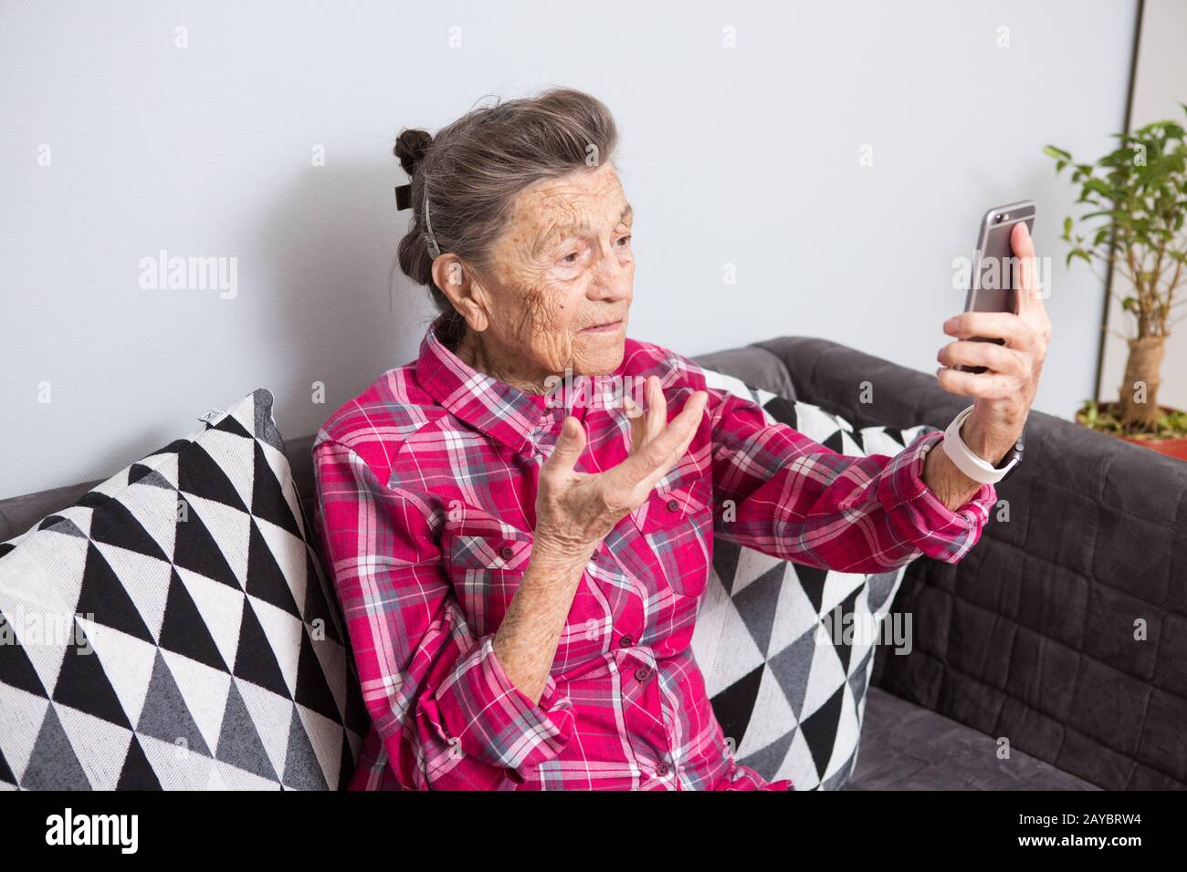 Una nonna caucasica molto vecchia con i capelli grigi e le rughe profonde siede a casa sul divano in jeans e camicia rossa a scacchi e. Foto Stock