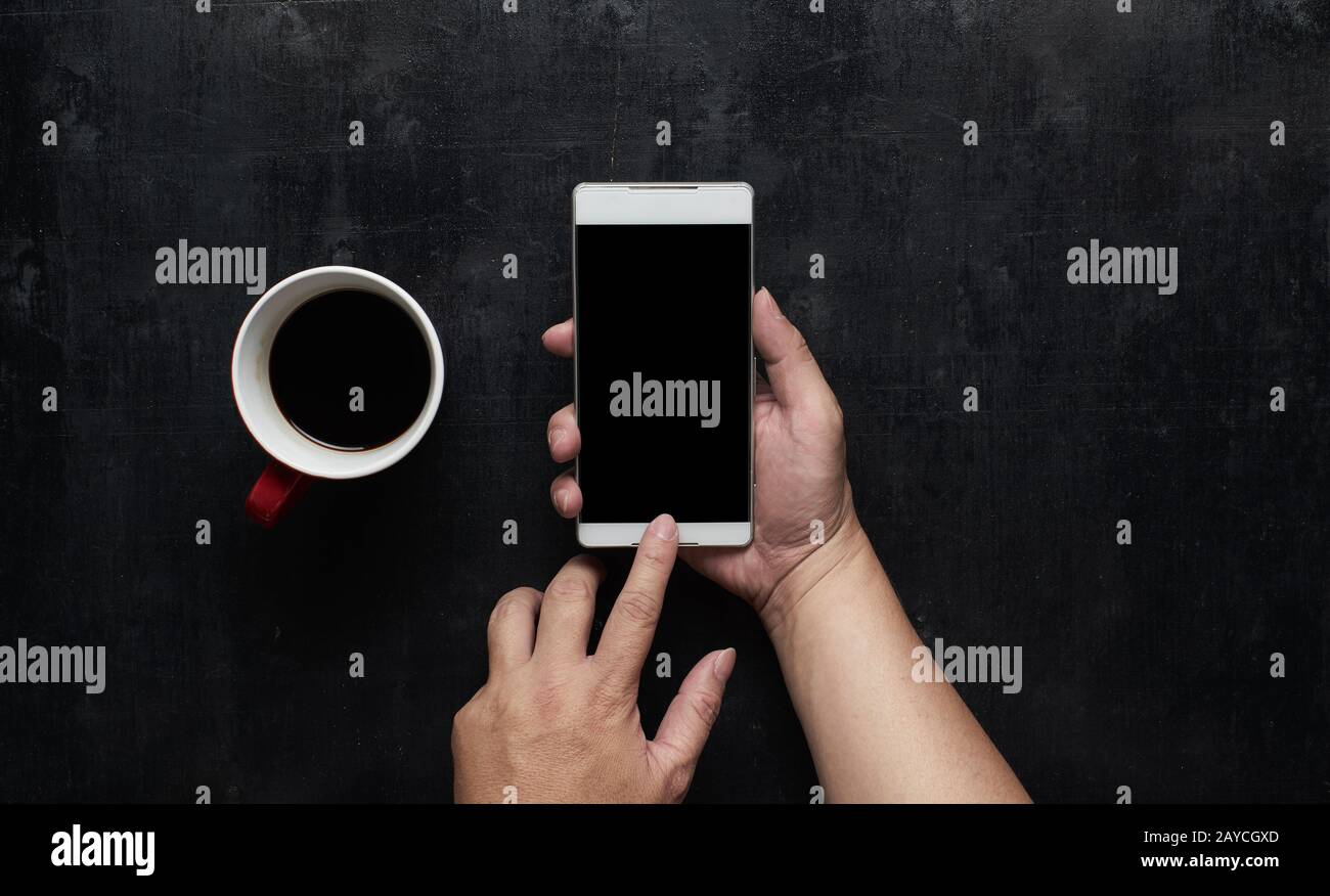 Mani che usano lo schermo nero del telefono con una tazza di caffè. Tavolo da ufficio nero in legno con vista dall'alto. Foto Stock