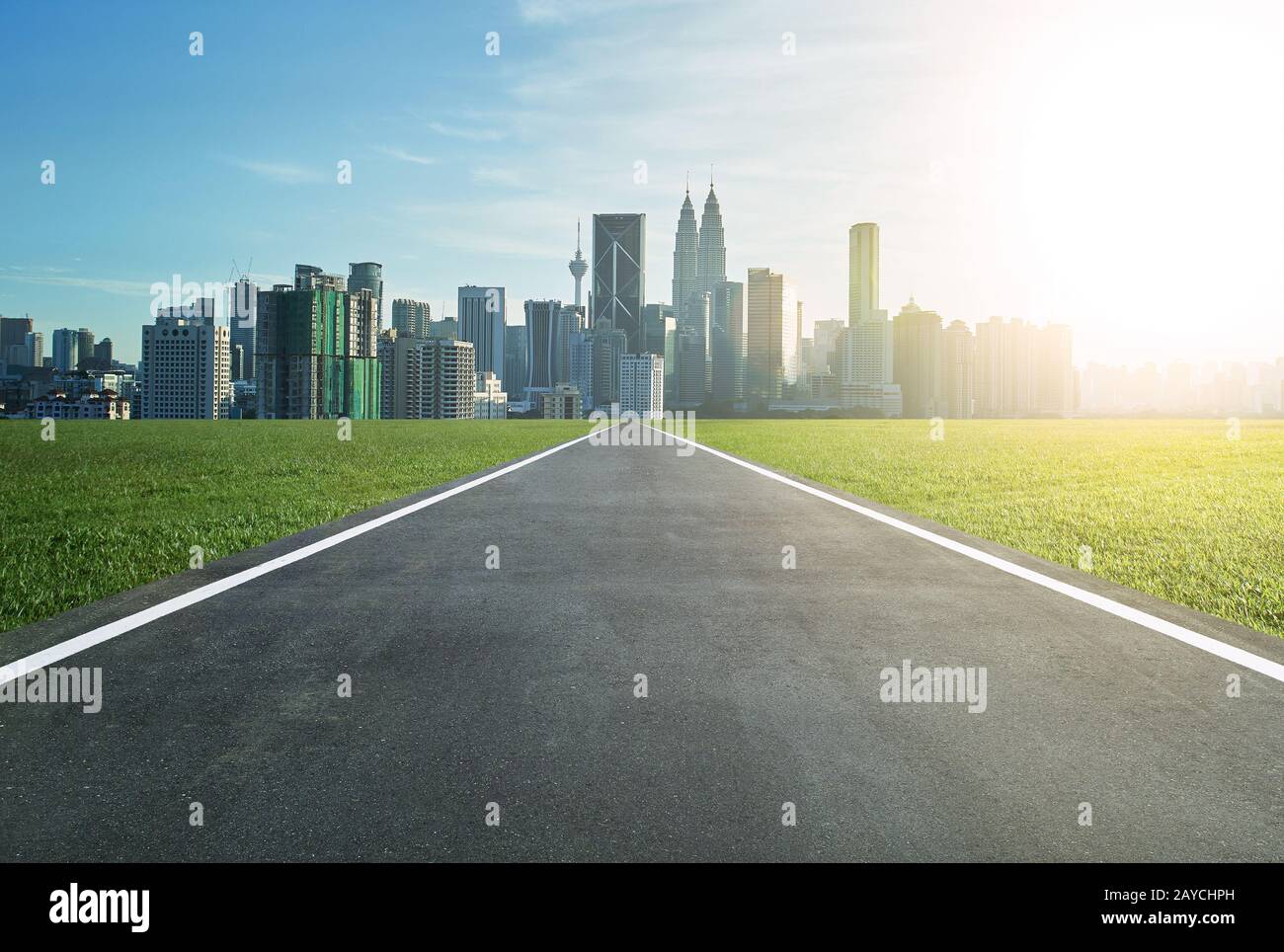 Strada asfaltata che conduce ad una città con alti edifici attraverso un prato verde Foto Stock