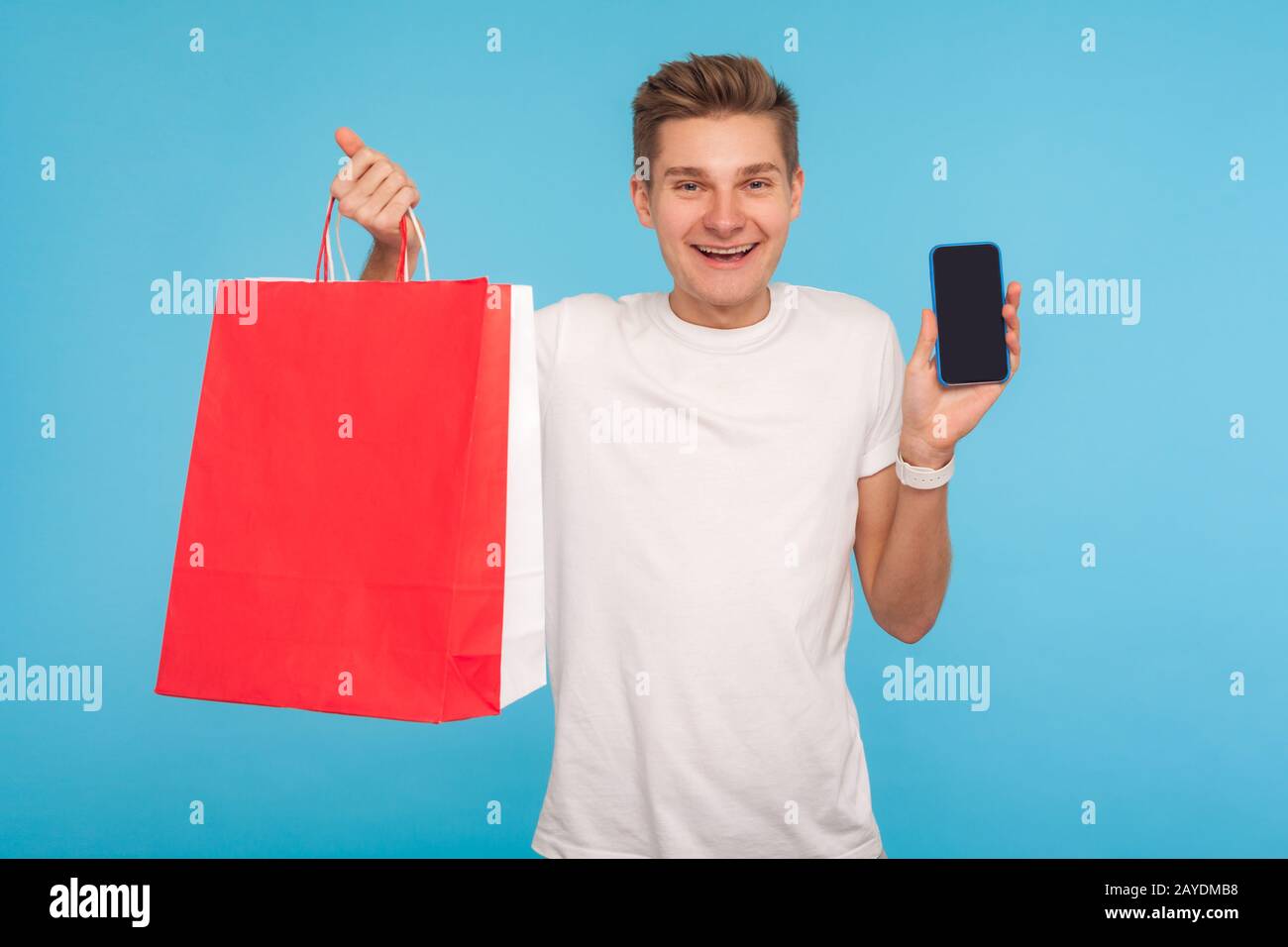 App mobile per lo shopping online. Gioioso uomo felice in casual t-shirt bianca tenendo cellulare e pacchetti, sorridendo alla fotocamera, pubblicità di sto online Foto Stock