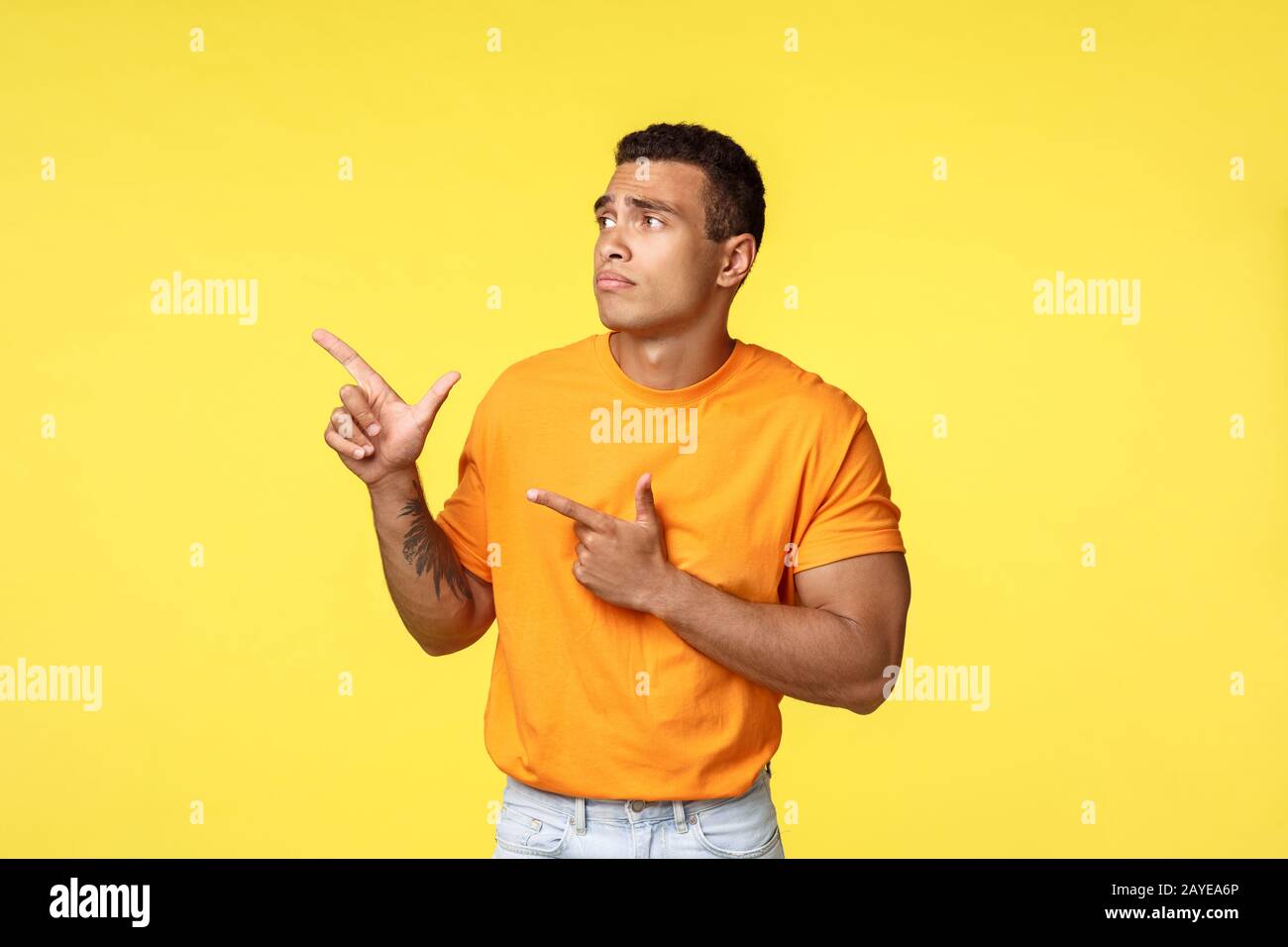 Geloso e sconvolto uomo carino maschile, indossare t-shirt arancione, in piedi infelice e triste su sfondo giallo, punto di spoloramento Foto Stock