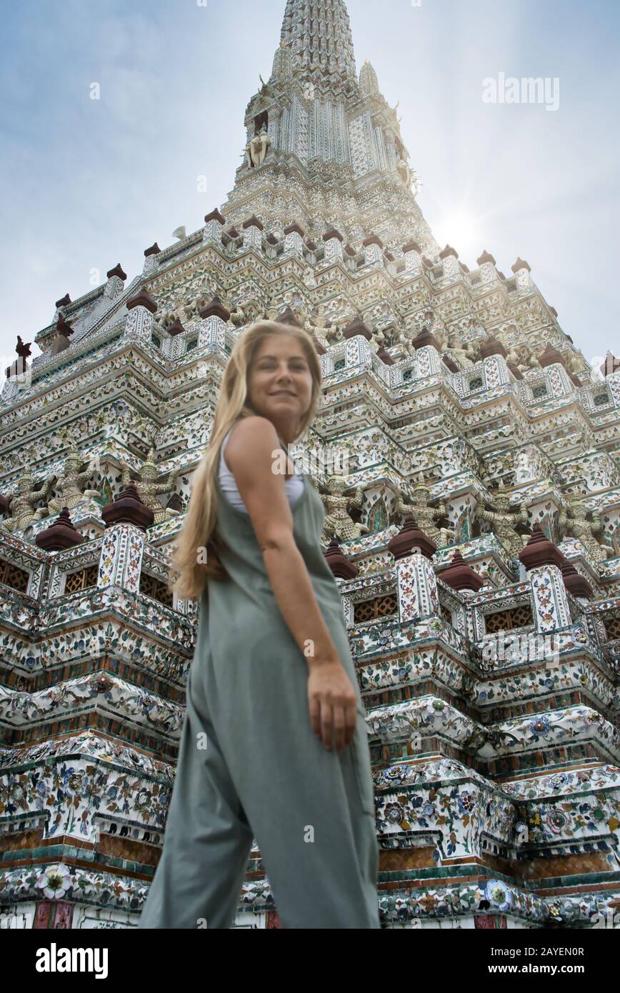 Foto verticale di una ragazza bionda che si erge davanti a un tempio buddista che gira intorno sorridendo in Thailandia Foto Stock
