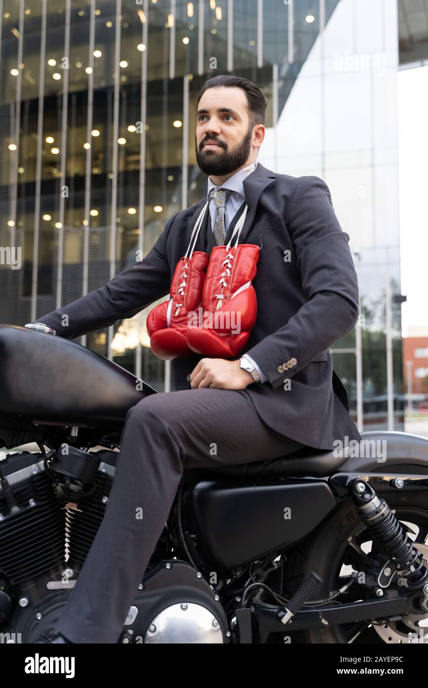 Uomo d'affari seduto su una motocicletta con guanti da boxe appesi al collo con alcuni edifici sullo sfondo Foto Stock