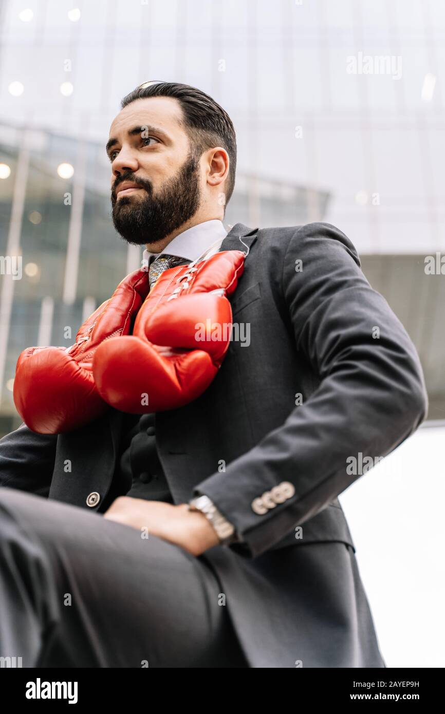 Foto verticale di un uomo d'affari seduto con guanti da boxe appeso al collo con alcuni edifici sullo sfondo Foto Stock