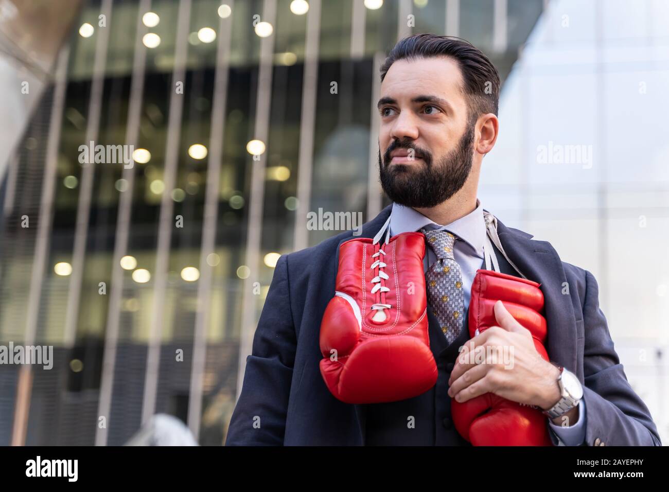 Uomo d'affari che indossa un vestito e che tocca i guanti di boxe rossi appesi dal collo davanti agli edifici Foto Stock