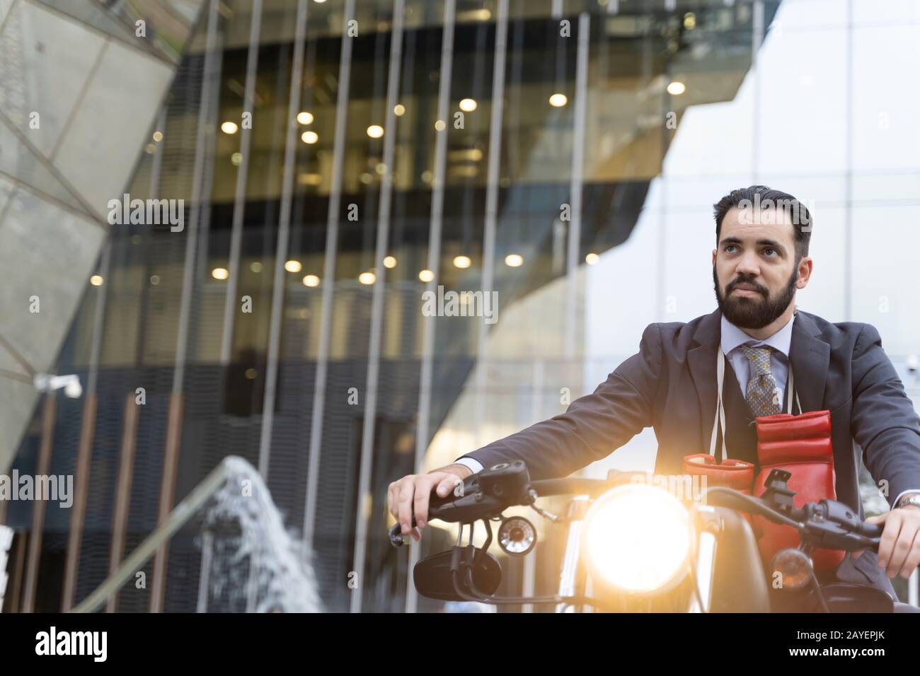 Uomo d'affari seduto su una moto con i guanti leggeri aperti e pugilato appeso al collo con alcuni edifici sullo sfondo Foto Stock