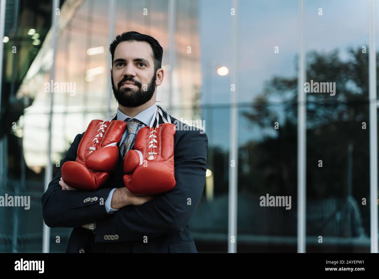 Uomo d'affari con guanti da boxe rossi appesi al collo con le braccia incrociate davanti a un edificio Foto Stock