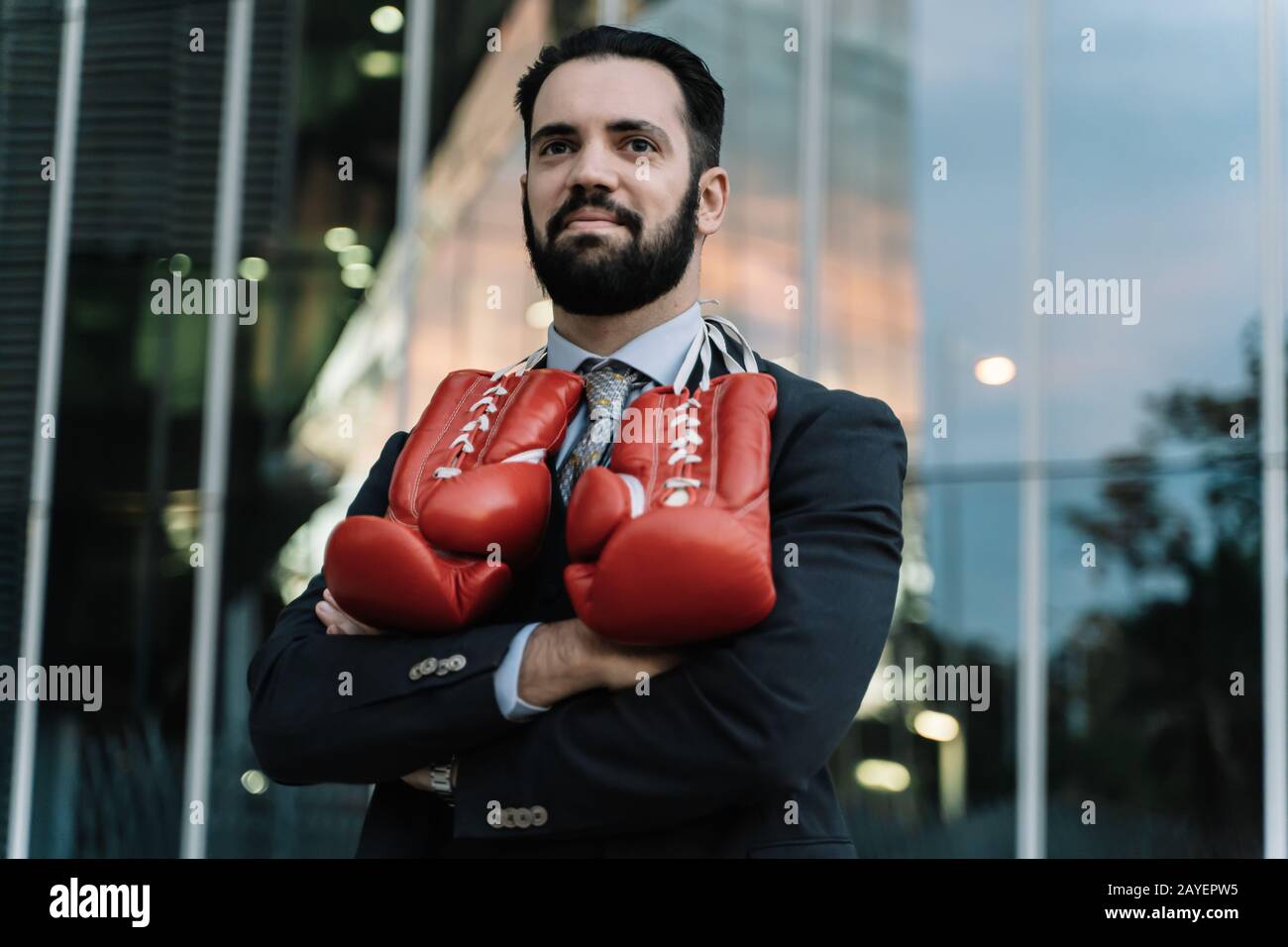 Uomo d'affari sorridente con guanti da boxe rossi appesi al collo con le braccia incrociate davanti a un edificio Foto Stock
