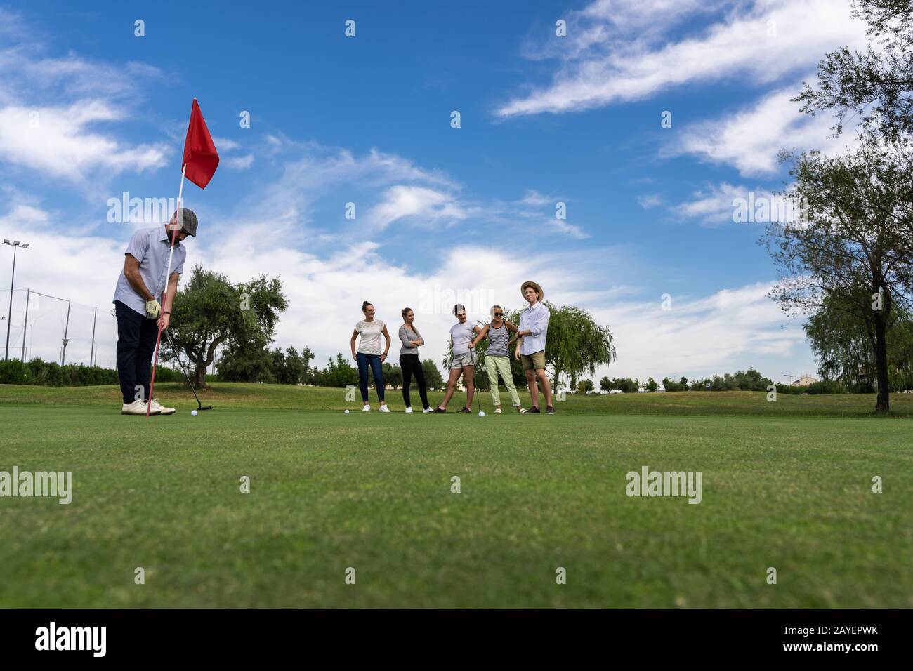 Uomo con un beretto che gioca a golf di fronte a un buco rosso-contrassegnato e un gruppo di persone che guardano su un campo da golf Foto Stock