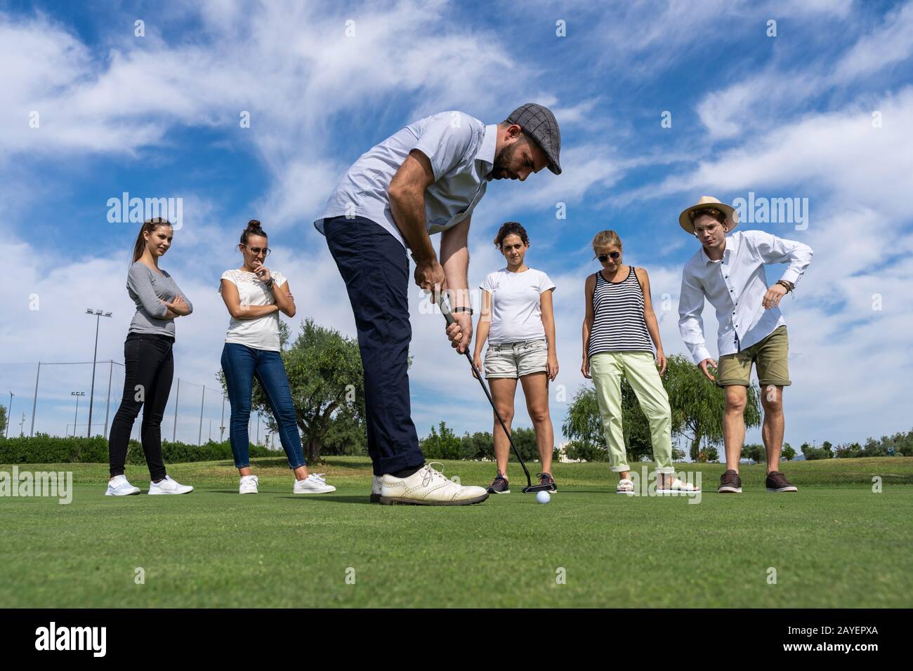 Giovane uomo con barba e beretto che gioca a golf con un golf club di fronte ad un gruppo di persone che lo guardano su un campo da golf Foto Stock