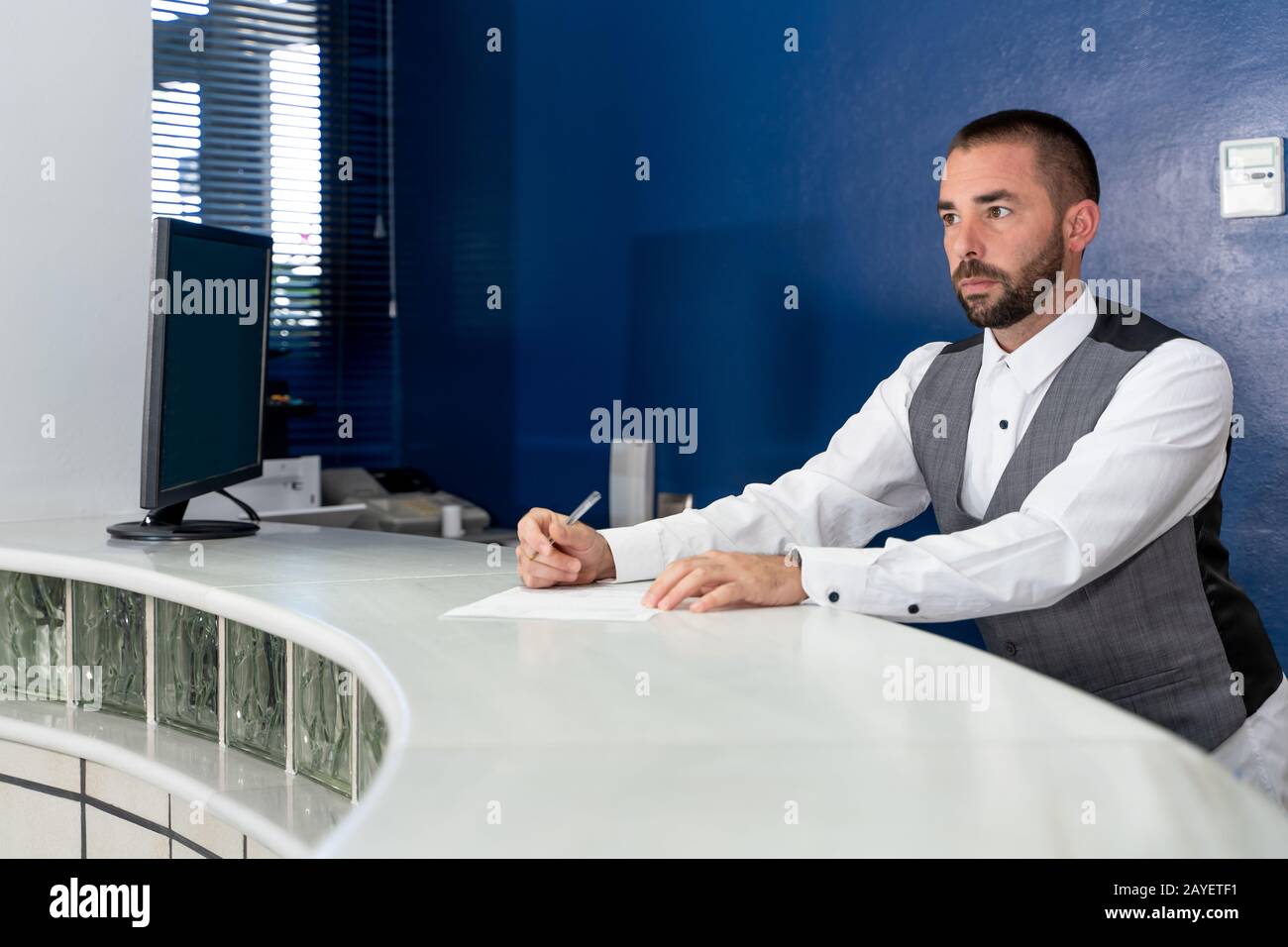 stock foto di un giovane uomo con mezza barba con camicia bianca e giubbotto che lavora in una reception dell'hotel Foto Stock