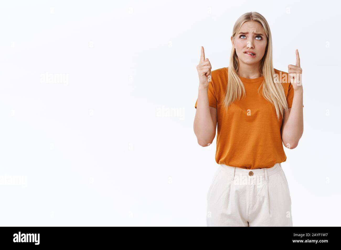 Preoccupato, giovane cute preoccupato donna bionda incerto in t-shirt arancione, pantaloni, labbro mordente ansioso, accigliato e guardando in alto, punta pubblicità superiore Foto Stock