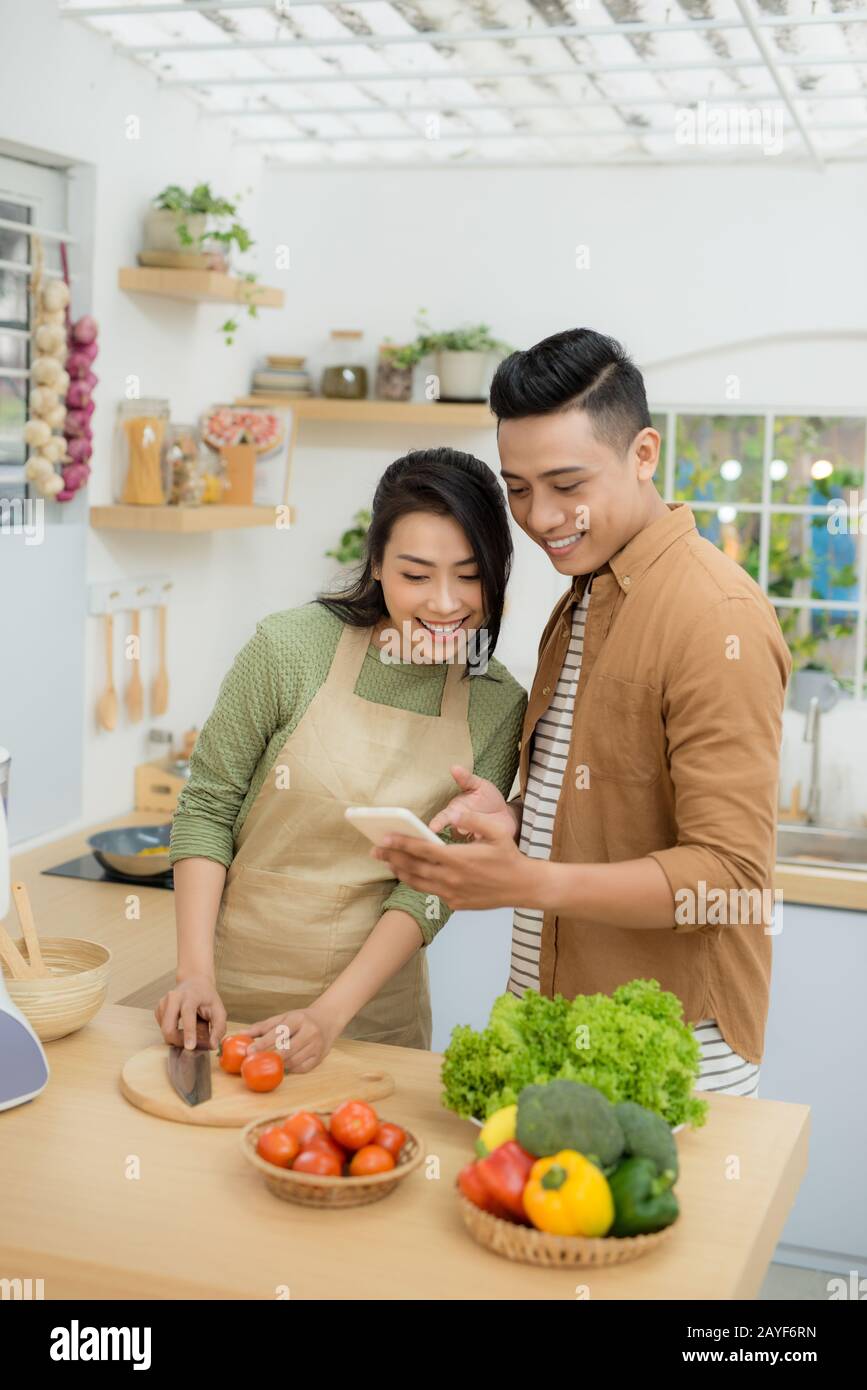 Bella coppia Giovane è utilizzando un telefono cellulare e sorridente durante la cottura in cucina a casa Foto Stock