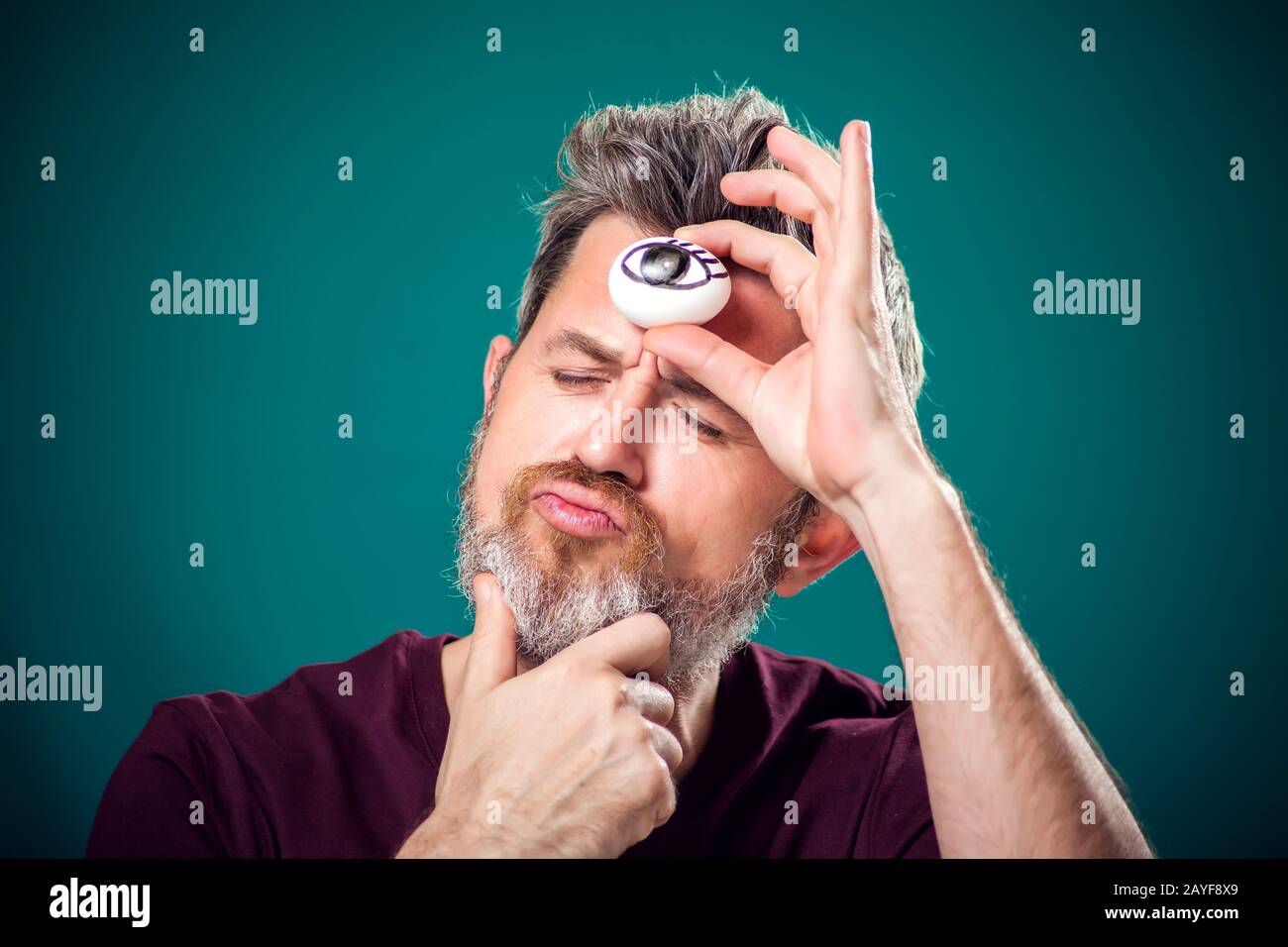 Un ritratto dell'uomo con bearded in t-shirt rossa che tiene l'uovo con l'occhio di disegno dietro forefad come terzo simbolo dell'occhio. Foto Stock