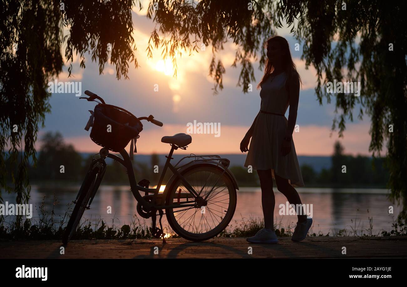Silhouette di donna in un abito e bicicletta retrò con un cesto sotto rami di albero sullo sfondo di un lago e il sole al tramonto. Magico paesaggio del tramonto Foto Stock