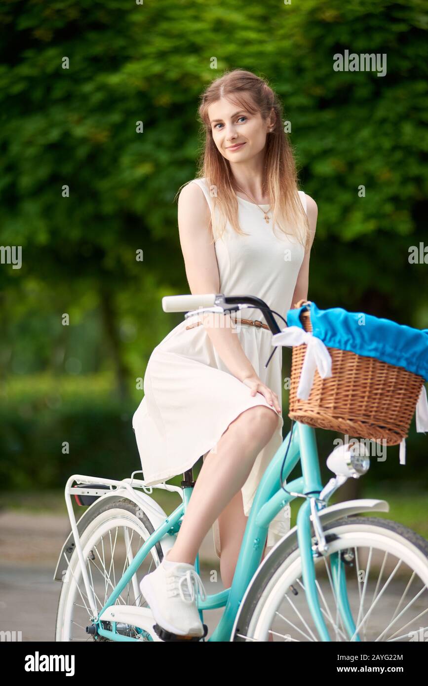 Ragazza carina in un abito leggero e sneakers bianche che posano mentre si siede su una bicicletta blu vintage con un cesto tra spazi verdi in un parco cittadino. Giorno fuori estate Foto Stock