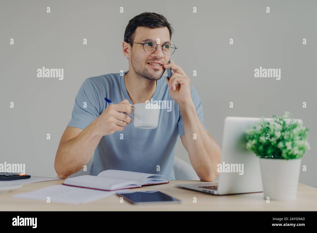 Piacevole aspetto contabile maschile in occhiali rotondi sips bevande calde, ha conversazione mobile, gestisce le finanze, funziona con computer portatile, non scrive Foto Stock